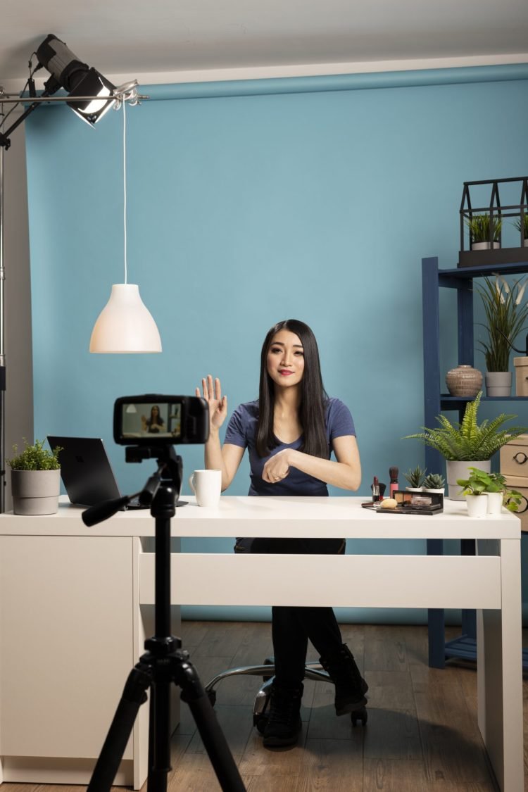 Beauty influencer sits at desk in a studio with a blue backdrop, engaging with her online audience during livestream. Perfect for themes of vlogging, content creation, and influencer marketing.