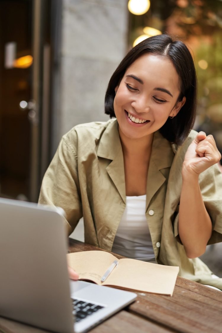 Vertical shot of happy girl talking on video call, looks at laptop, having online meeting, sitting in outdoor cafe.